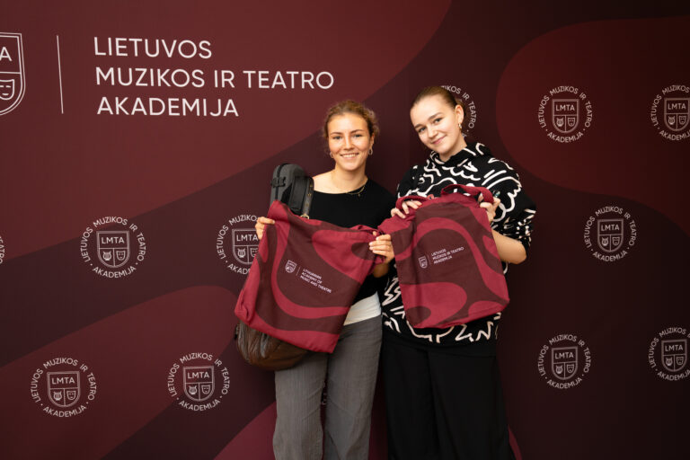 Du jaunos moterys pozuoja prie sienos su logotipais, laikydamos tamsiai raudonus pirkinių maišelius su LMTA logotipu.
