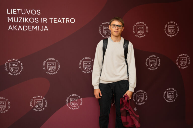 Jaunas vyras su akiniais ir kuprine stovi prieš rudą foną su baltu užrašu "LIETUVOS MUZIKOS IR TEATRO AKADEMIJA" ir besikartojančiu akademijos logotipu.
