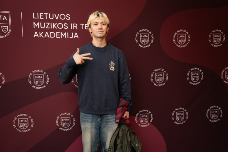 Jaunas vyras su šviesiais plaukais daro taikos gestą prieš tamsiai raudoną foną su baltu logotipu.