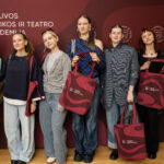 Septyni jauni žmonės stovi kartu prieš tamsiai raudoną foną su logotipu, ant kurio užrašyta "Lietuvos muzikos ir teatro akademija". Jie visi laiko bordo spalvos pirkinių krepšius su tokia pačia akademijos logotipu.