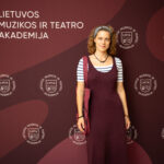 Moteris, apsirengusi bordo spalvos suknelę ir dryžuotus marškinėlius, stovi prieš tamsiai raudoną foną su baltu logotipu ir užrašu "LIETUVOS MUZIKOS IR TEATRO AKADEMIJA".