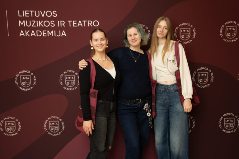 Trys moterys stovi prieš tamsiai raudoną foną su logotipais, ant kurio užrašyta "LIETUVOS MUZIKOS IR TEATRO AKADEMIJA".