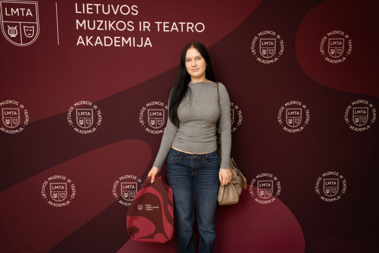 Jauna moteris stovi prieš raudoną foną su logotipais ir užrašu "Lietuvos muzikos ir teatro akademija", laiko rankoje raudoną maišelį ir ant peties turi rankinę.