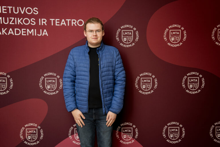 Jaunas vyras su akiniais ir mėlynu pūkinuku stovi prieš raudonų fonų su užrašais ir logotipais.