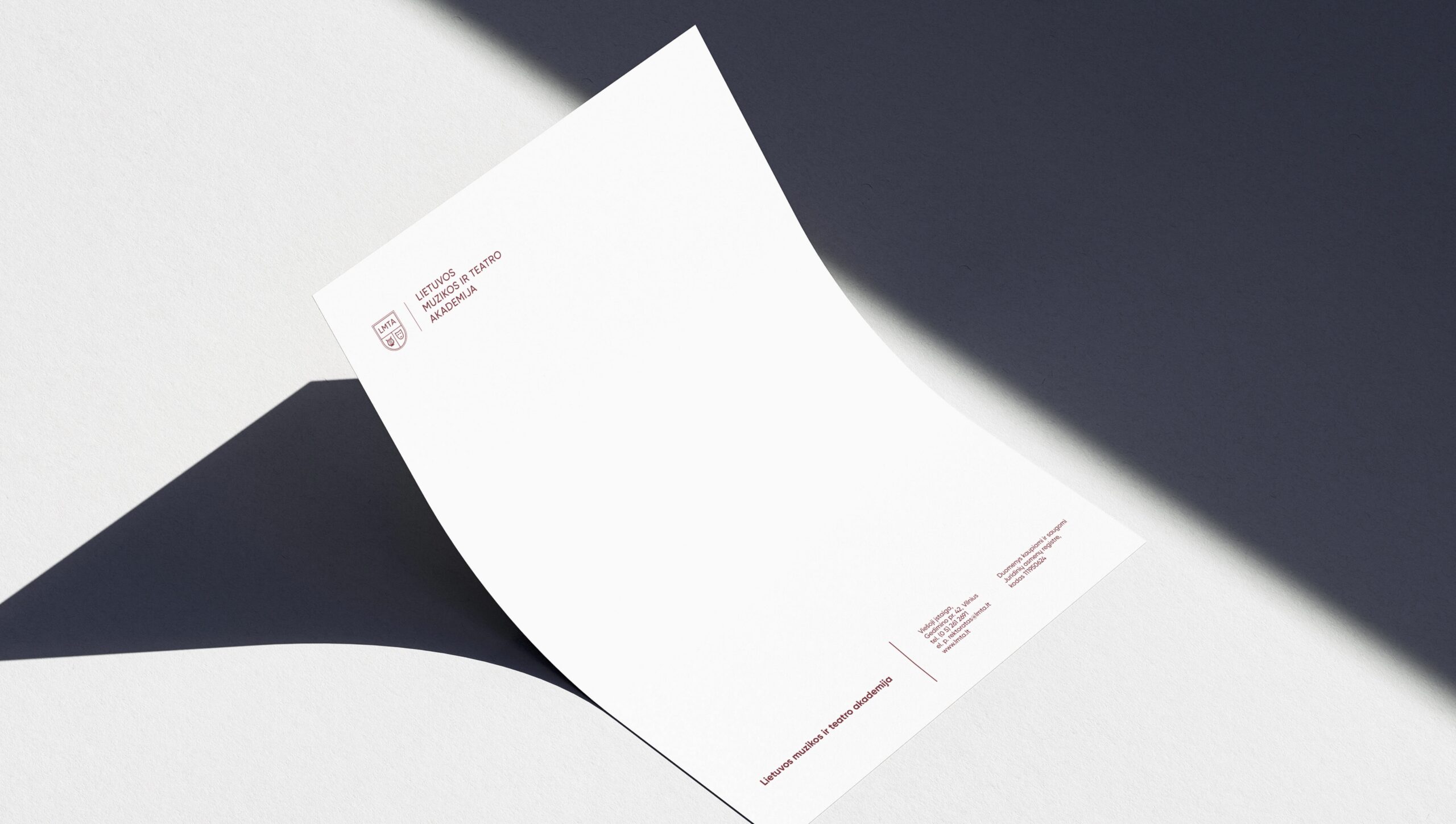 Balta popieriaus lapo dalis, užversta ant tamsaus paviršiaus, su raudonu logotipu ir tekstu „Lietuvos muzikos ir teatro akademija“ viename kampe, bei kontaktine informacija kitame.