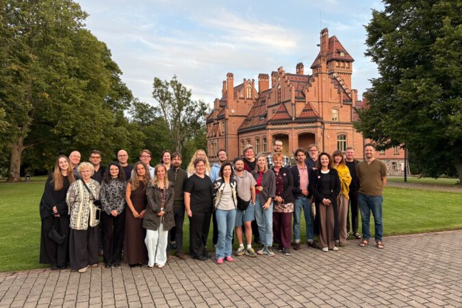 Draugų grupė pozuoja prie plytinio dviaukščio dvaro, apsupto žalių medžių.