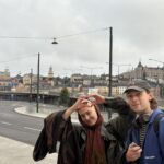 Poros, besisvečiuojančios Stokholme, atlieka širdies formą rankomis su miesto peizažu fone.