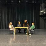 Keturi aktoriai repeticijose scenoje ant grindų, apsupti juodų užuolaidų, sėdi aplink geltoną stalą.