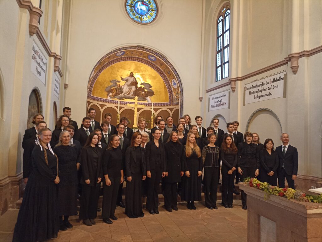 Didelė choro grupė, apsirengusi juodais koncertiniais drabužiais, pozuoja bažnyčioje priešais auksinę mozaikinę apsidę su Kristaus atvaizdu.