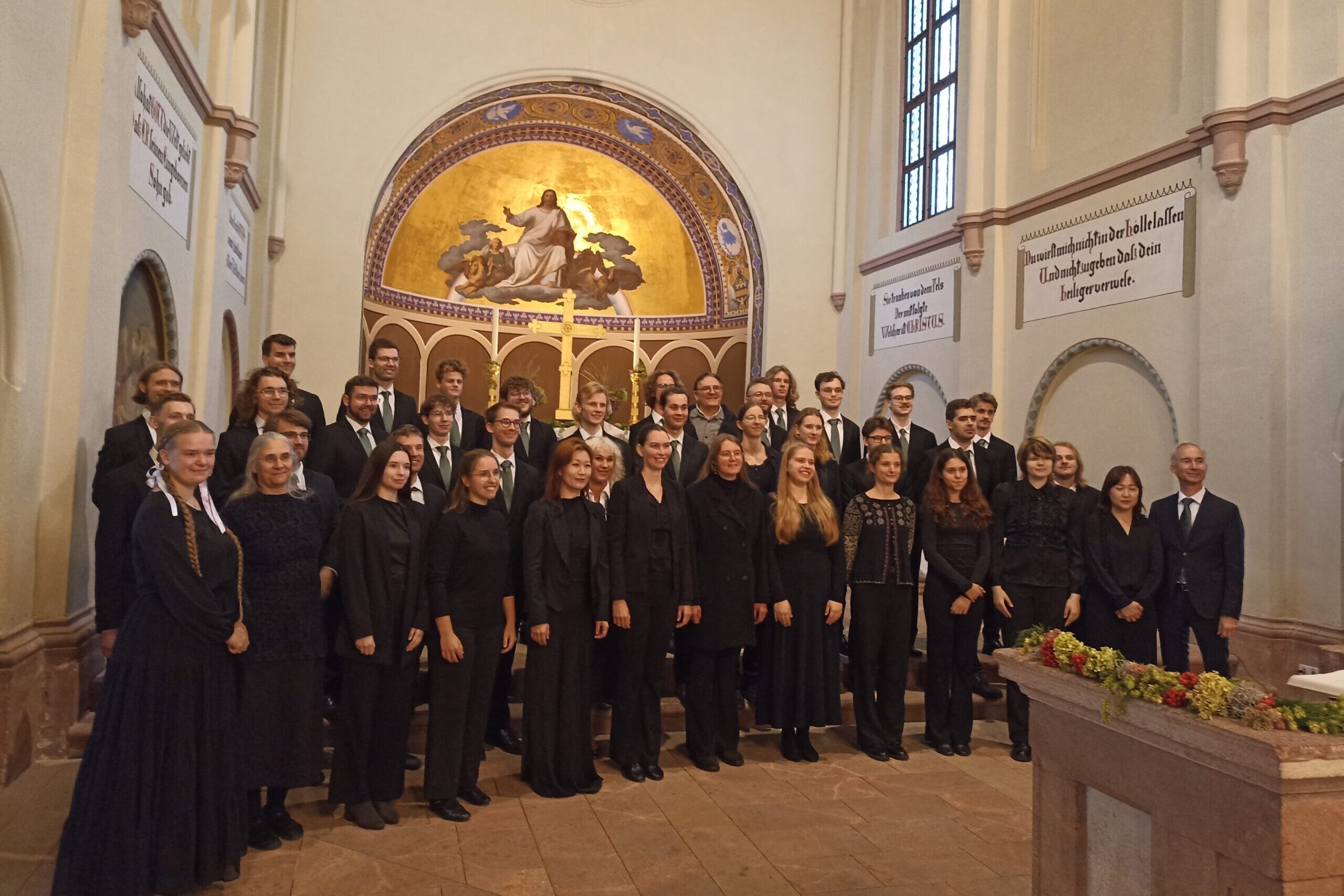Didelė choro grupė, apsirengusi juodais koncertiniais drabužiais, pozuoja bažnyčioje priešais auksinę mozaikinę apsidę su Kristaus atvaizdu.