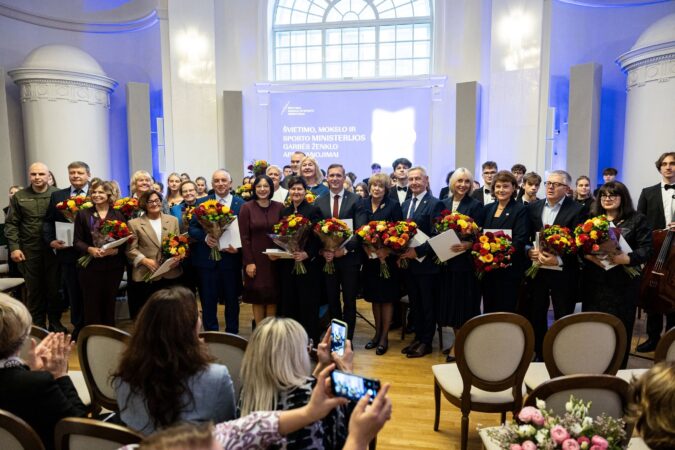 Švietimo, mokslo ir sporto ministerijos Garbės ženklo apdovanojimų ceremonijos laureatai su gėlių puokštėmis ir diplomais stovi salėje bendrai nuotraukai, o publika juos fotografuoja.