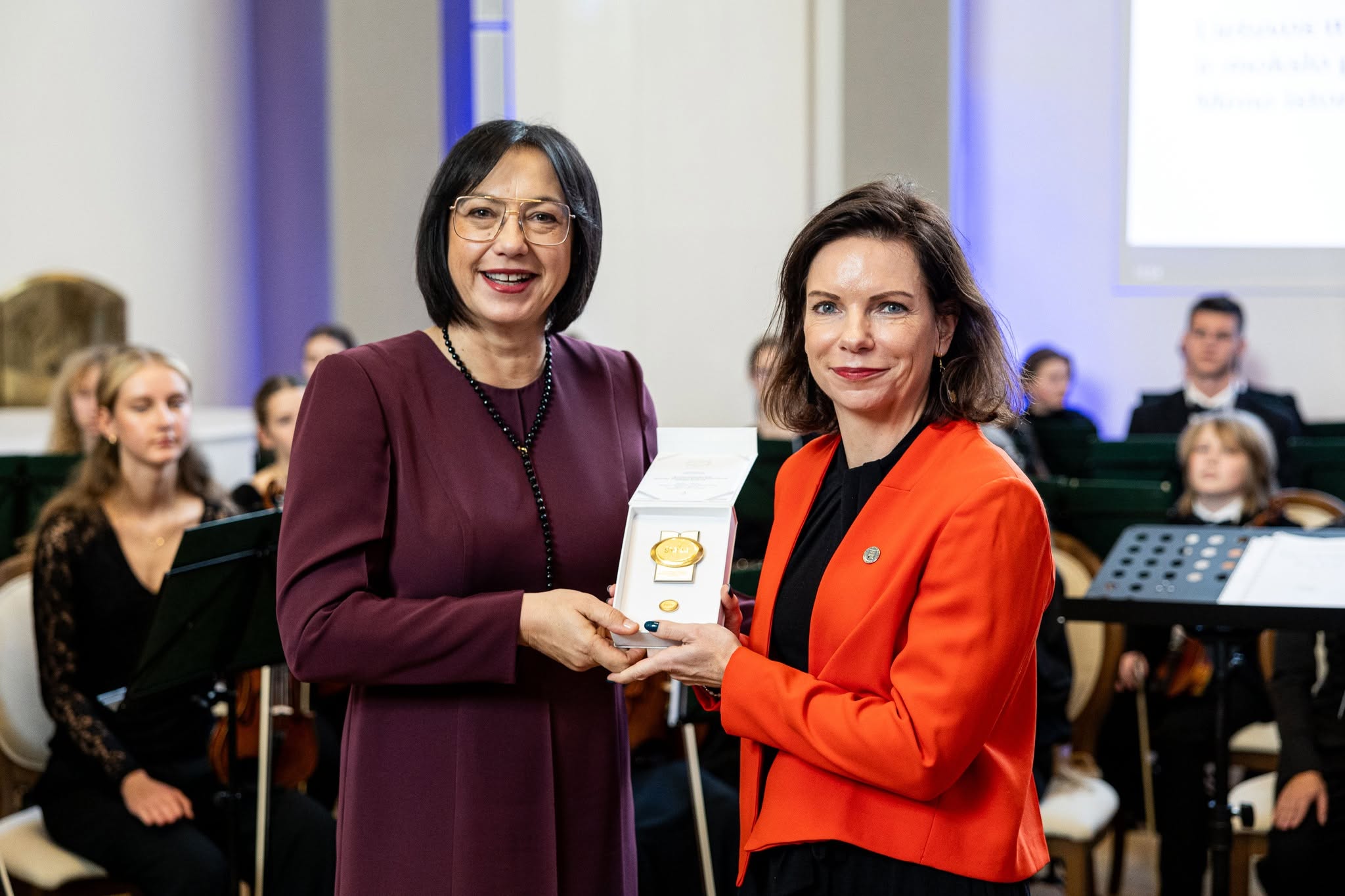 Dvi besišypsančios moterys laiko atidarytą dėžutę su auksiniu medaliu orkestro fone oficialaus renginio metu.