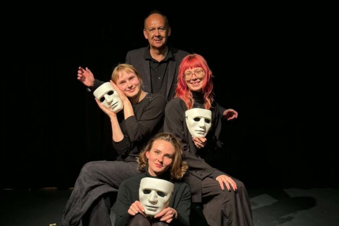 Keturi juodai apsirengę aktoriai tamsioje scenoje: trys moterys priekyje laiko baltas teatro kaukes, o už jų stovi vyras.