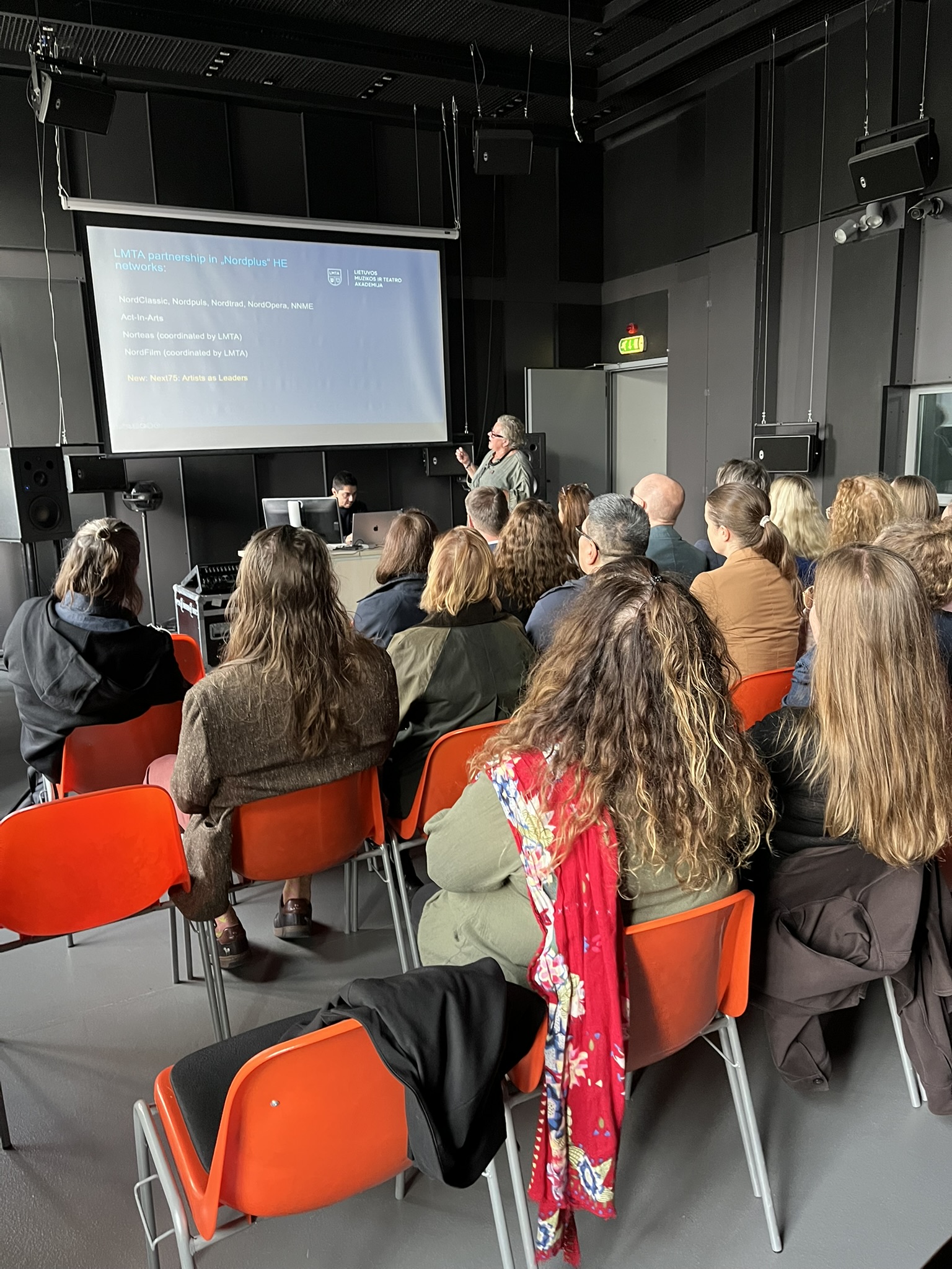 Žmonių grupė sėdi auditorijoje ant oranžinių kėdžių ir klausosi pranešėjos, rodančios į skaidrę projektoriaus ekrane.