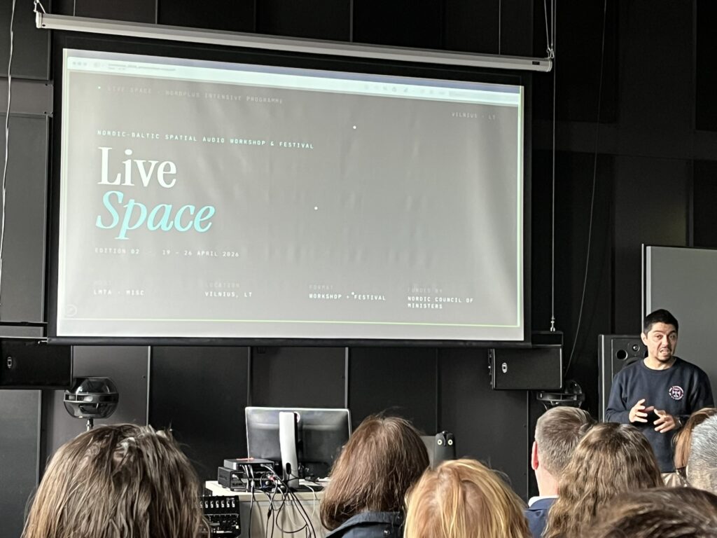 Pranešėjas stovi šalia didelio ekrano su skaidre apie „Live Space“ renginį Vilniuje priešais susirinkusius klausytojus.