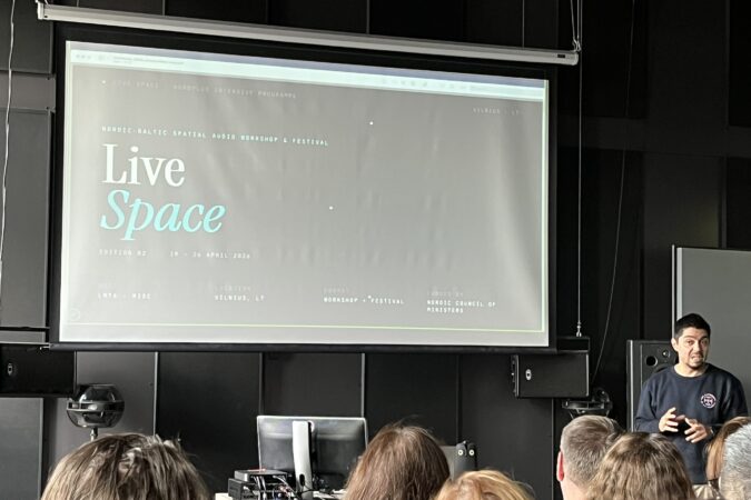 Pranešėjas stovi šalia didelio ekrano su skaidre apie „Live Space“ renginį Vilniuje priešais susirinkusius klausytojus.
