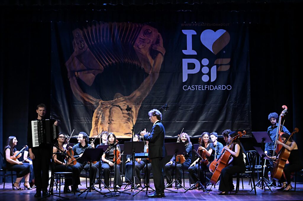 Styginių orkestras su dirigentu ir akordeonistu scenoje priešais „I Love PIF Castelfidardo“ reklamjuostę.