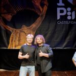 Du besišypsantys jauni vyrai scenoje rodo pakeltus nykščius tamsaus renginio plakato su akordeono atvaizdu fone.