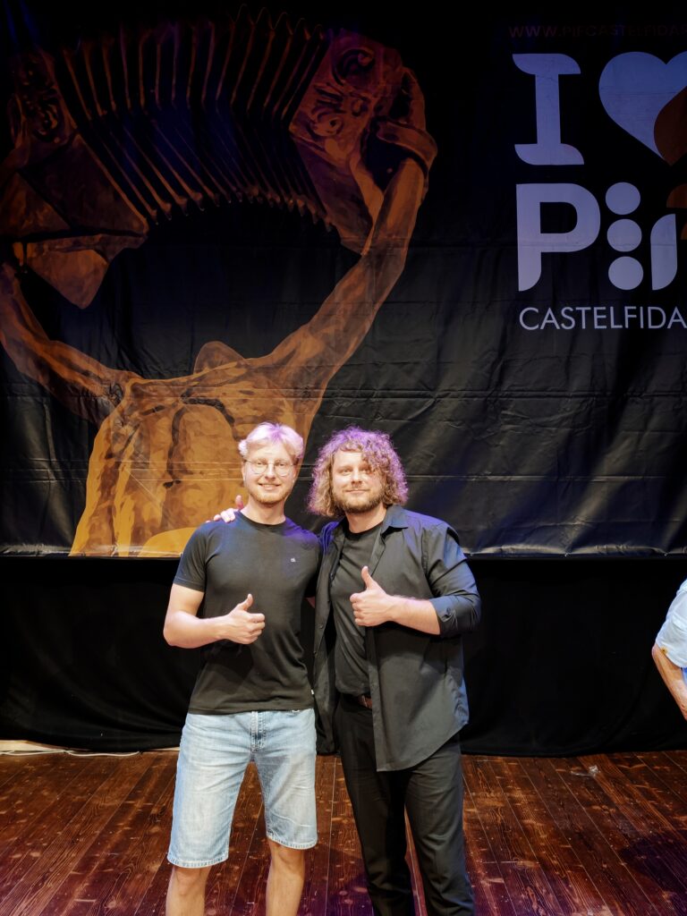 Du besišypsantys jauni vyrai scenoje rodo pakeltus nykščius tamsaus renginio plakato su akordeono atvaizdu fone.